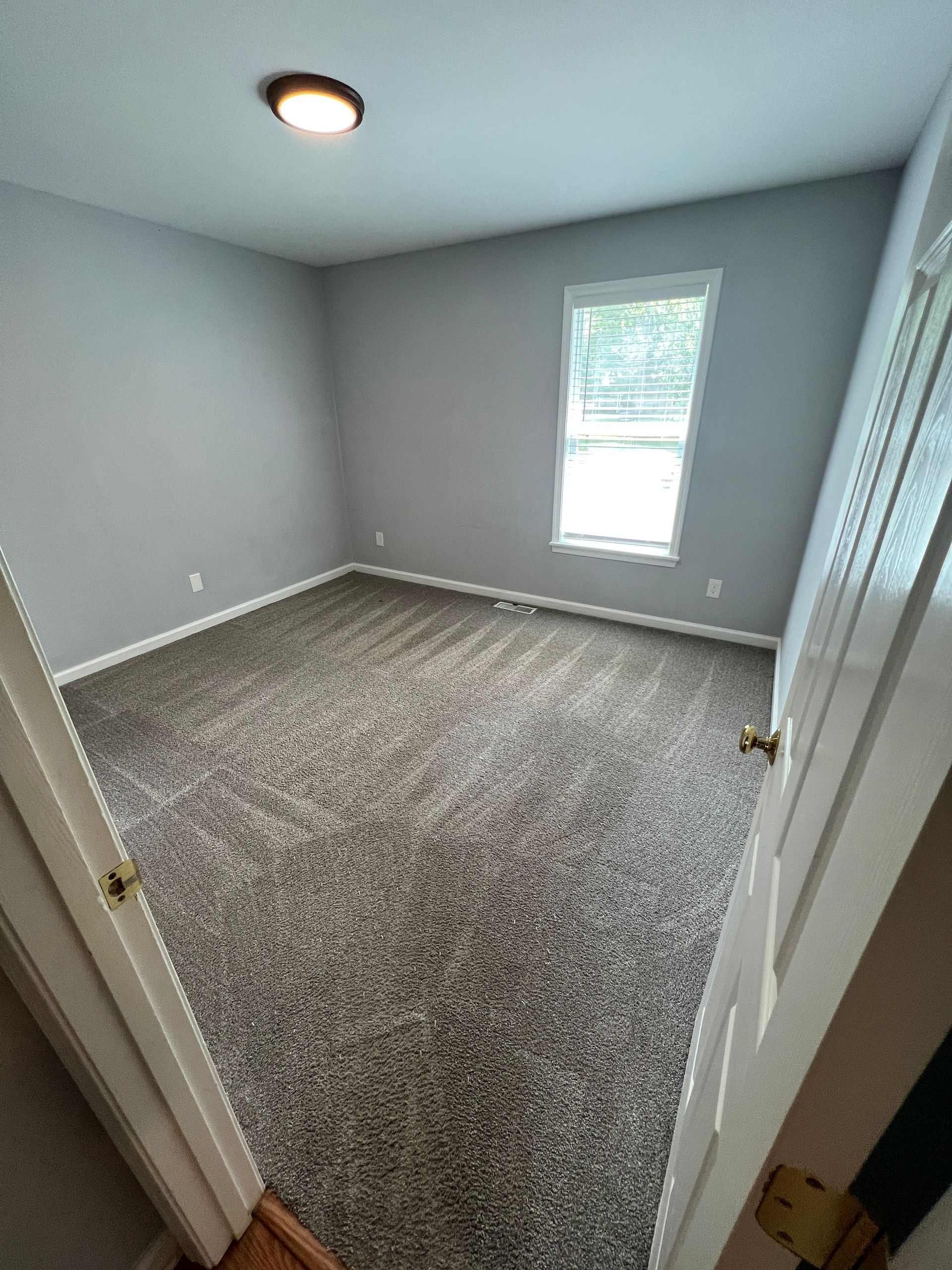 Empty gray-carpeted room with light gray walls, ceiling light, and a single window.