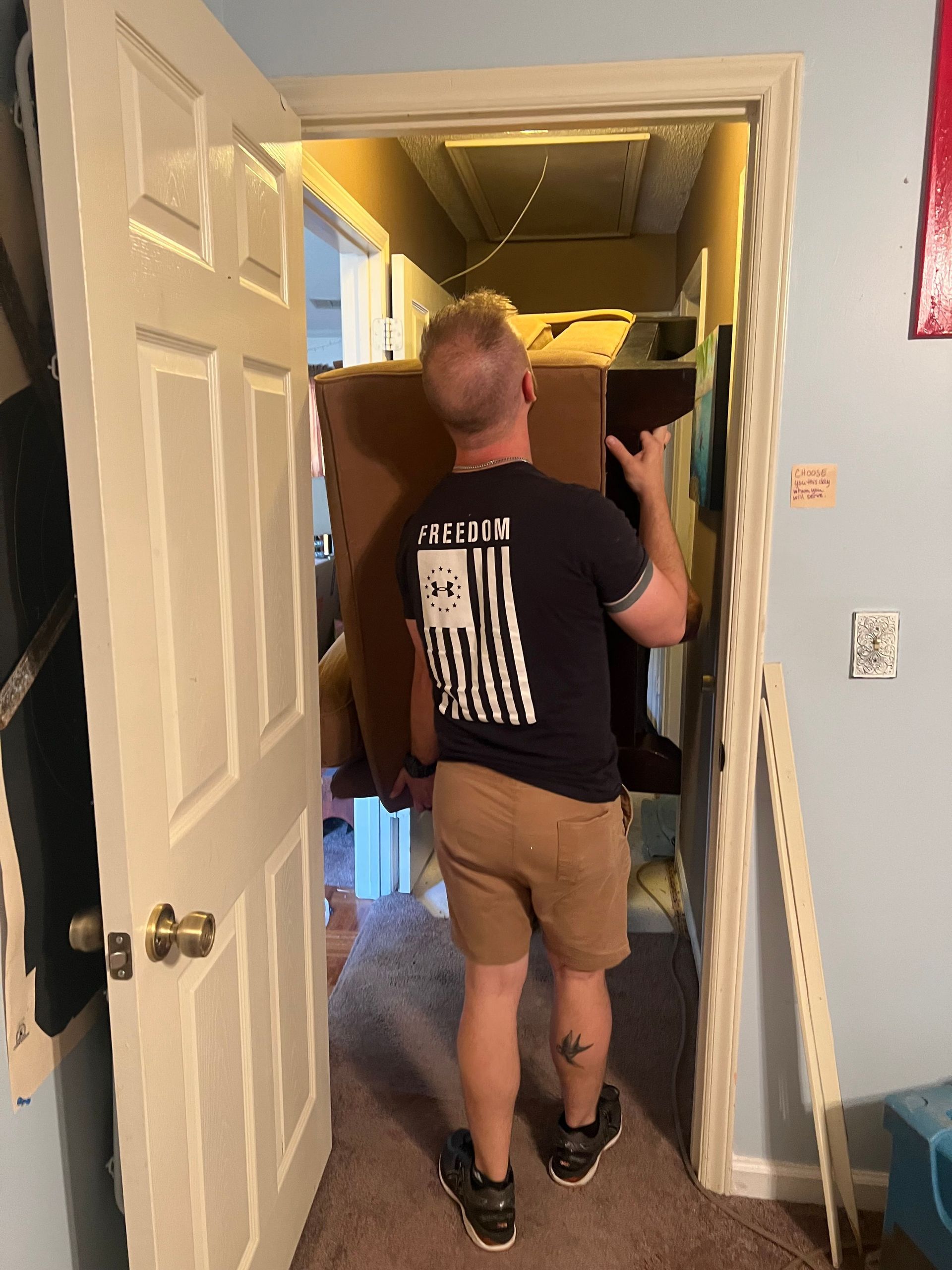 Person standing in a doorway, carrying a yellow object down a narrow hallway with a staircase railing.