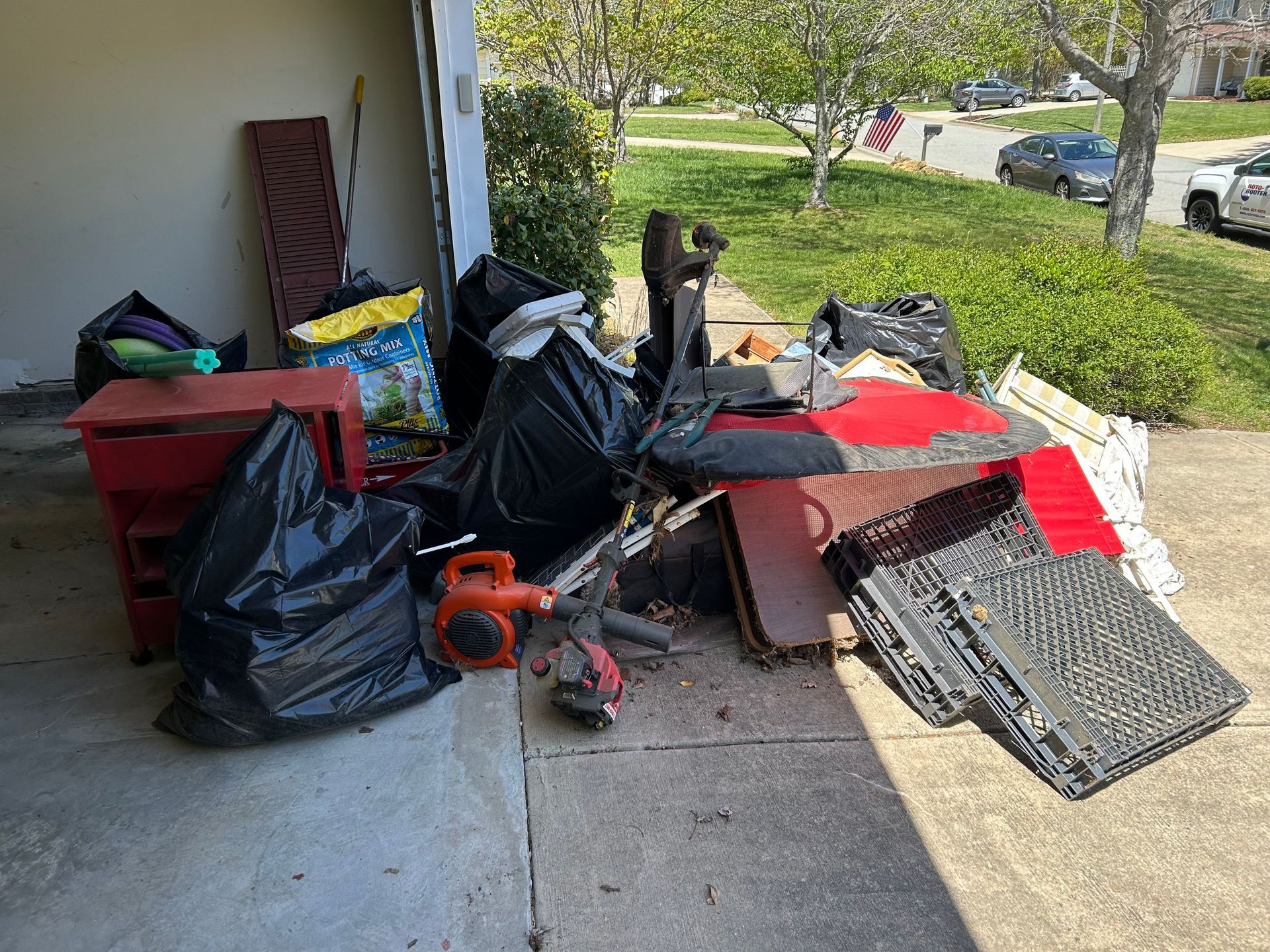 Pile of household junk and debris on a porch and driveway beside a house, including bins, bags, boards, and a leaf blower.