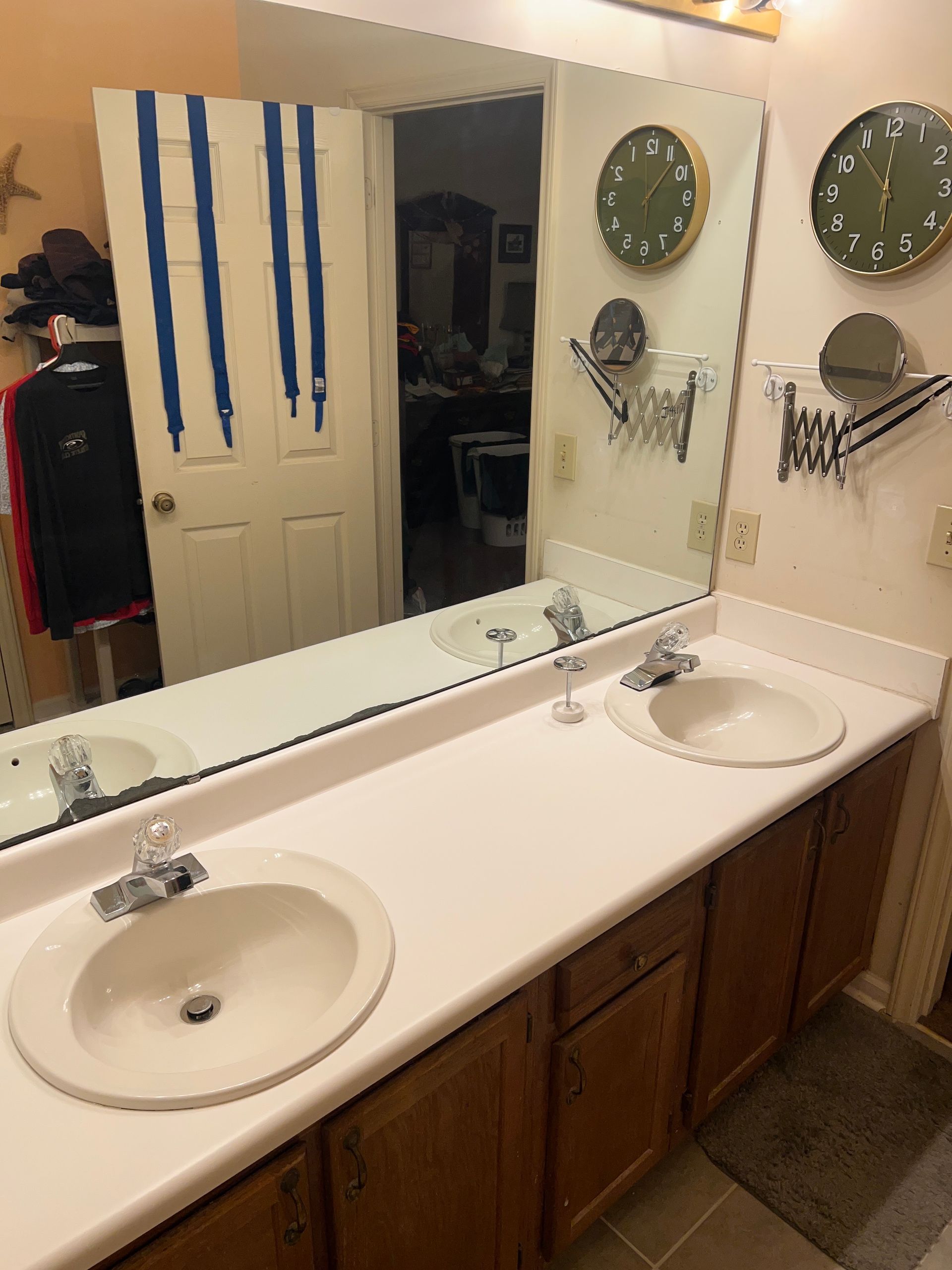 Bathroom vanity with two sinks, large mirror, green wall clocks, and blue-striped towels hanging on the door.
