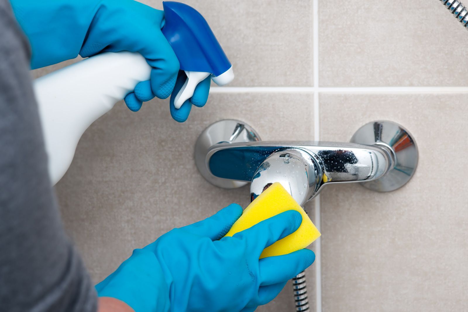 Gloved hands cleaning a chrome shower faucet with a yellow sponge in a tiled bathroom.