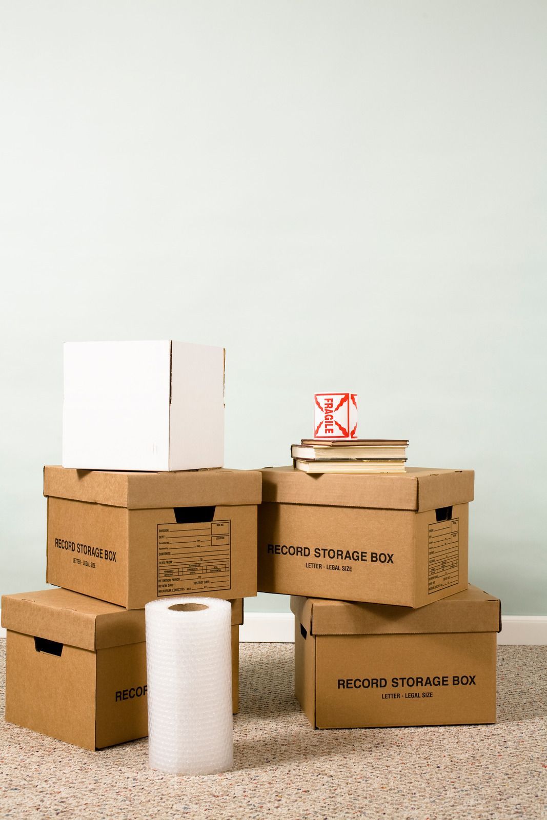 Stacked moving boxes on carpet, with a roll of bubble wrap in front and a blank wall behind.