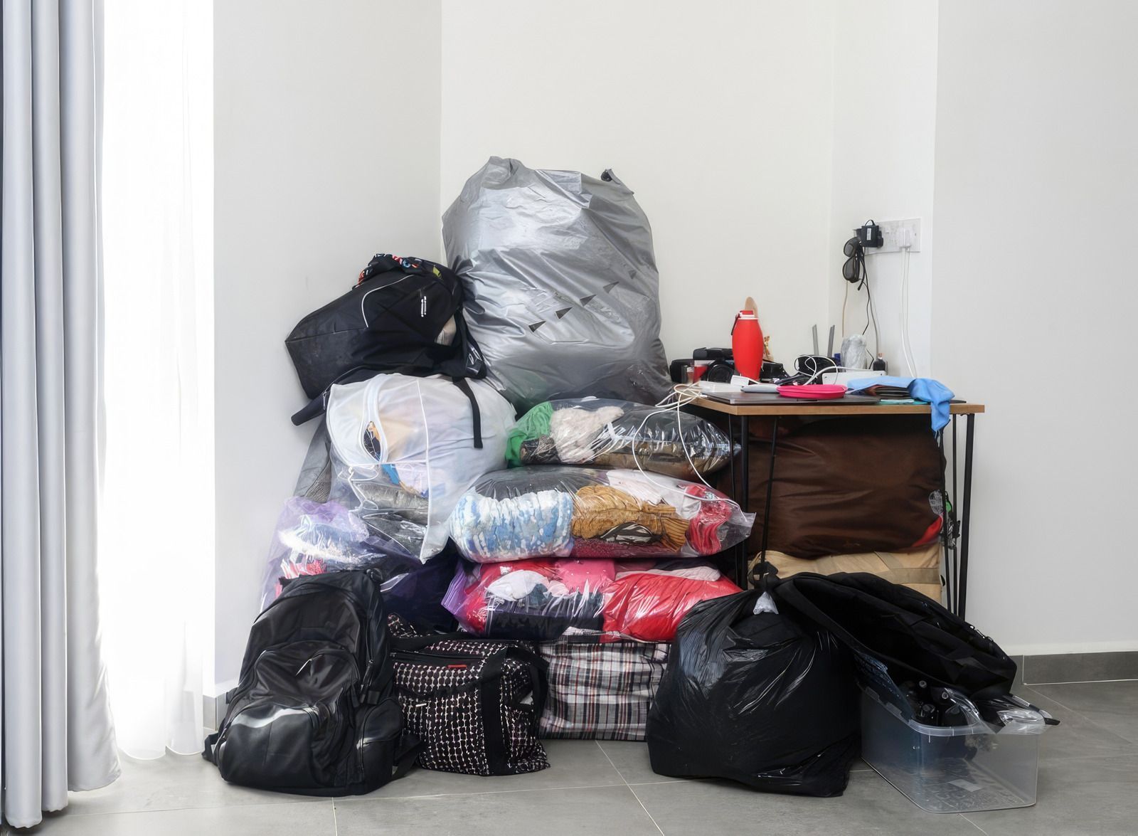 Piled bags and folded blankets stacked against a wall beside a small table with items on top