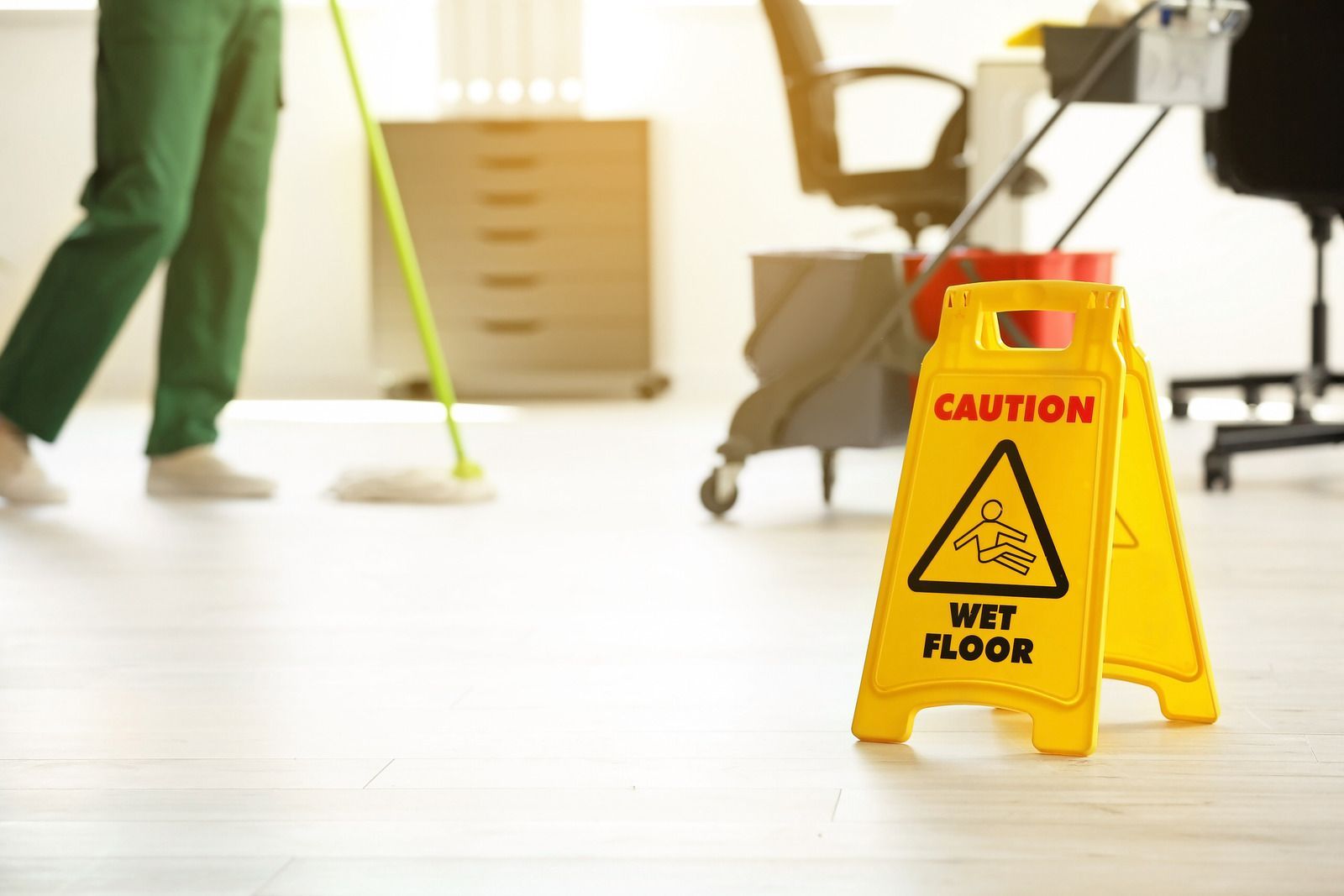 Caution wet floor sign on a bright office hallway floor, with a person mopping in the background