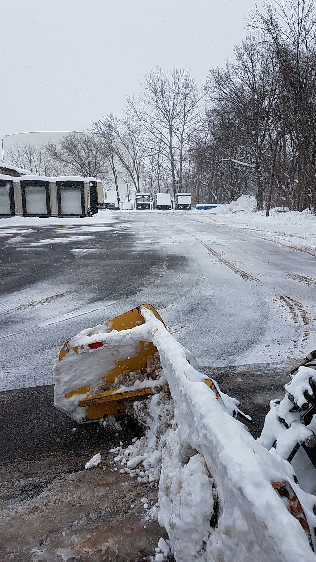 Yellow snowplow clearing a snow-covered parking area with loading docks in the background on a snowy day.