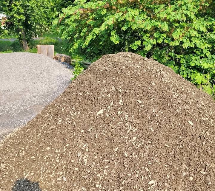 Pile of dark mulch in foreground, with a pile of gravel and trees in the background.