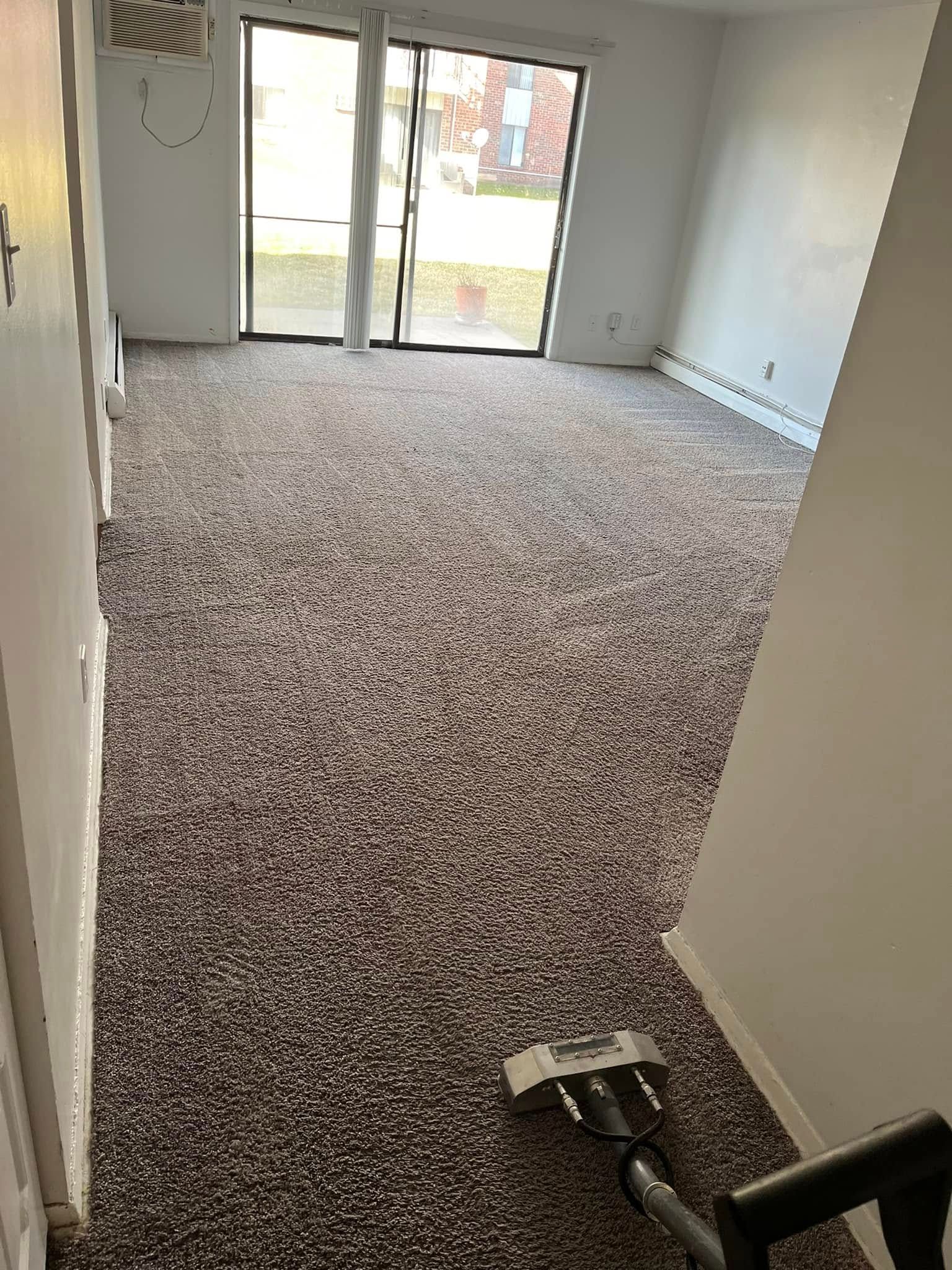 Empty carpeted room with sliding glass doors, white walls, and a vacuum cleaner in the foreground
