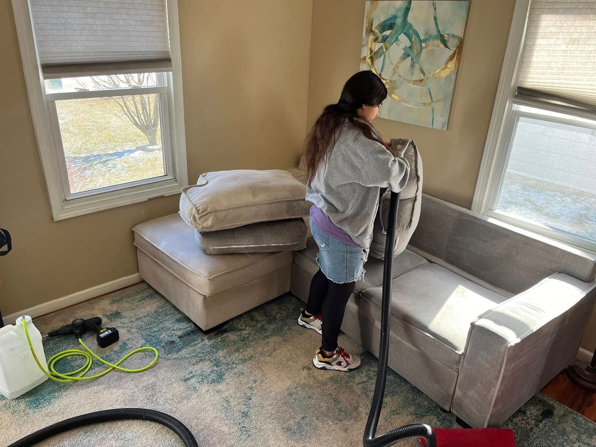 Person vacuuming a small living room with a gray couch, bed, and windows.