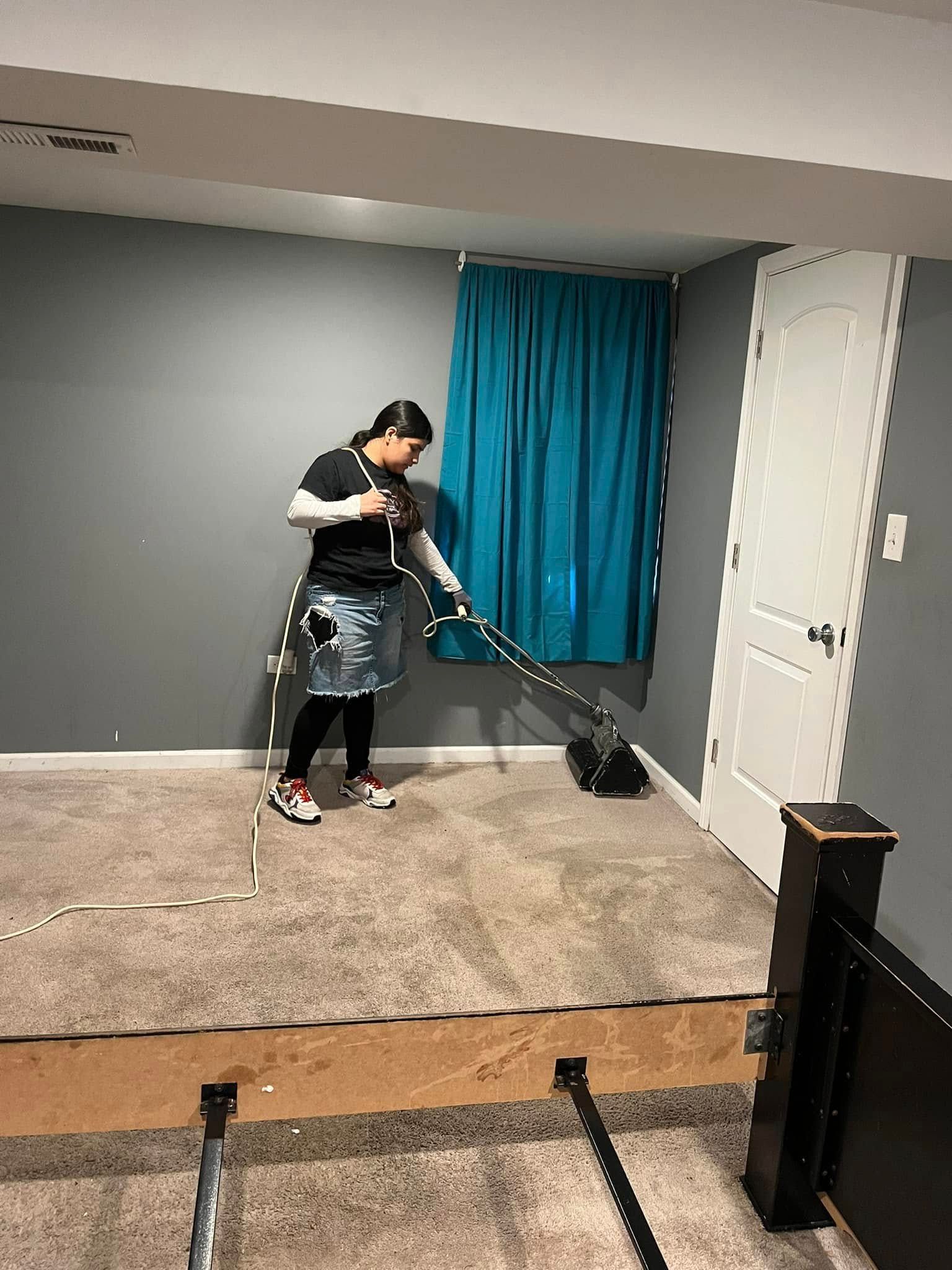 Person vacuuming a beige carpet in a room with gray walls and blue curtains.