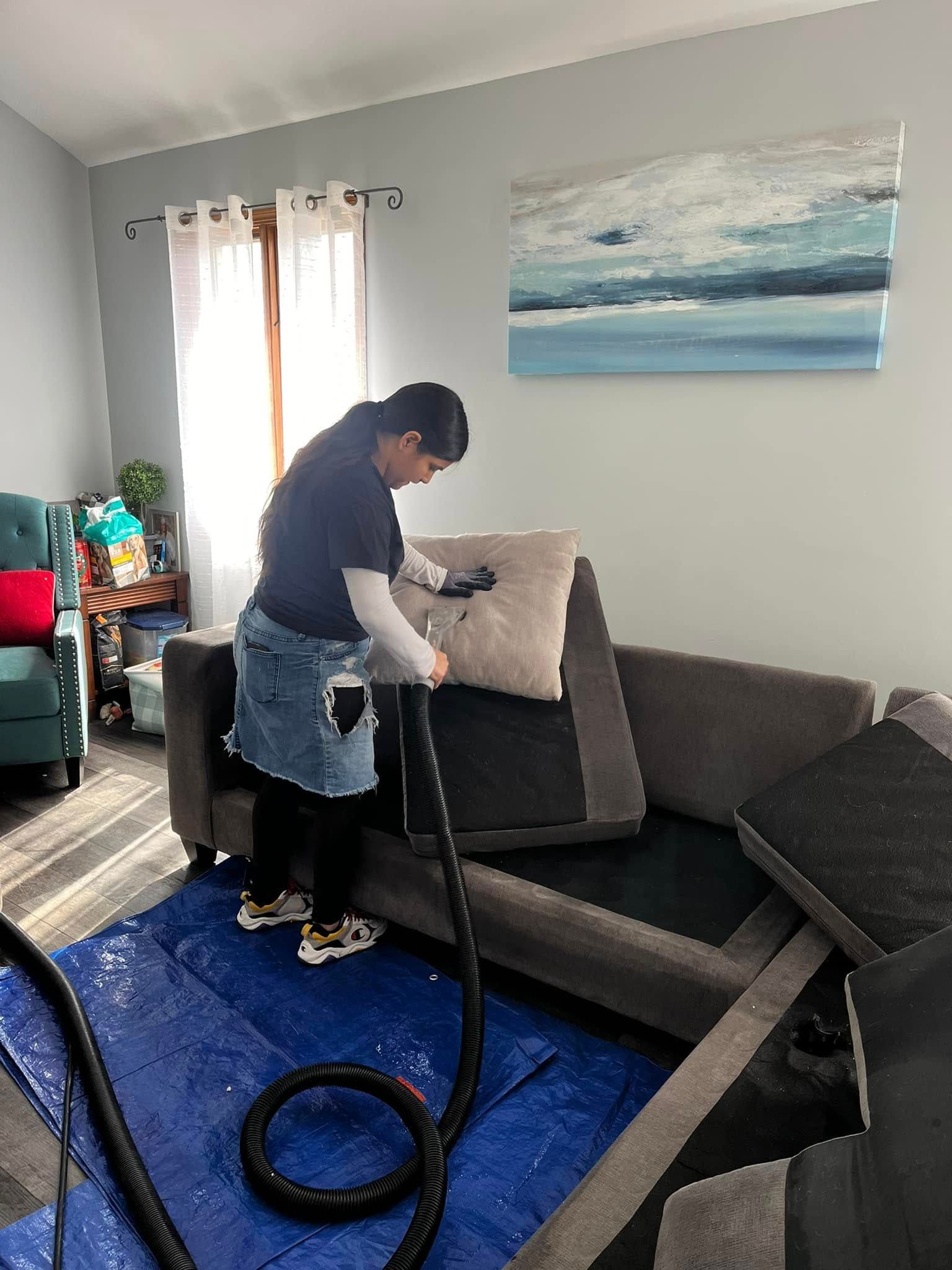 Person vacuuming a gray couch in a bright living room with a blue rug and wall art.