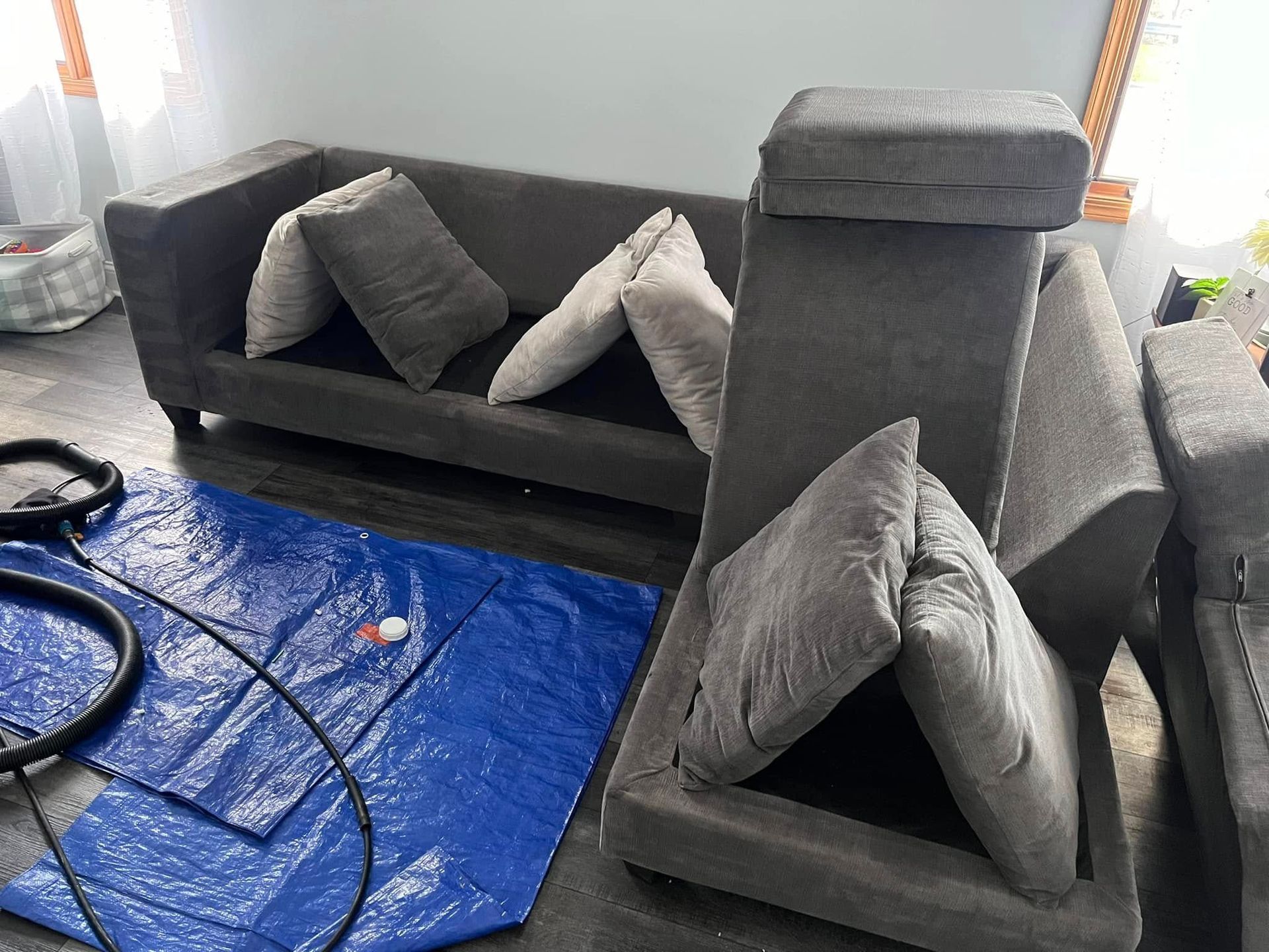 Gray couch cushions and pillows arranged in a room, with a blue tarp on the floor nearby.