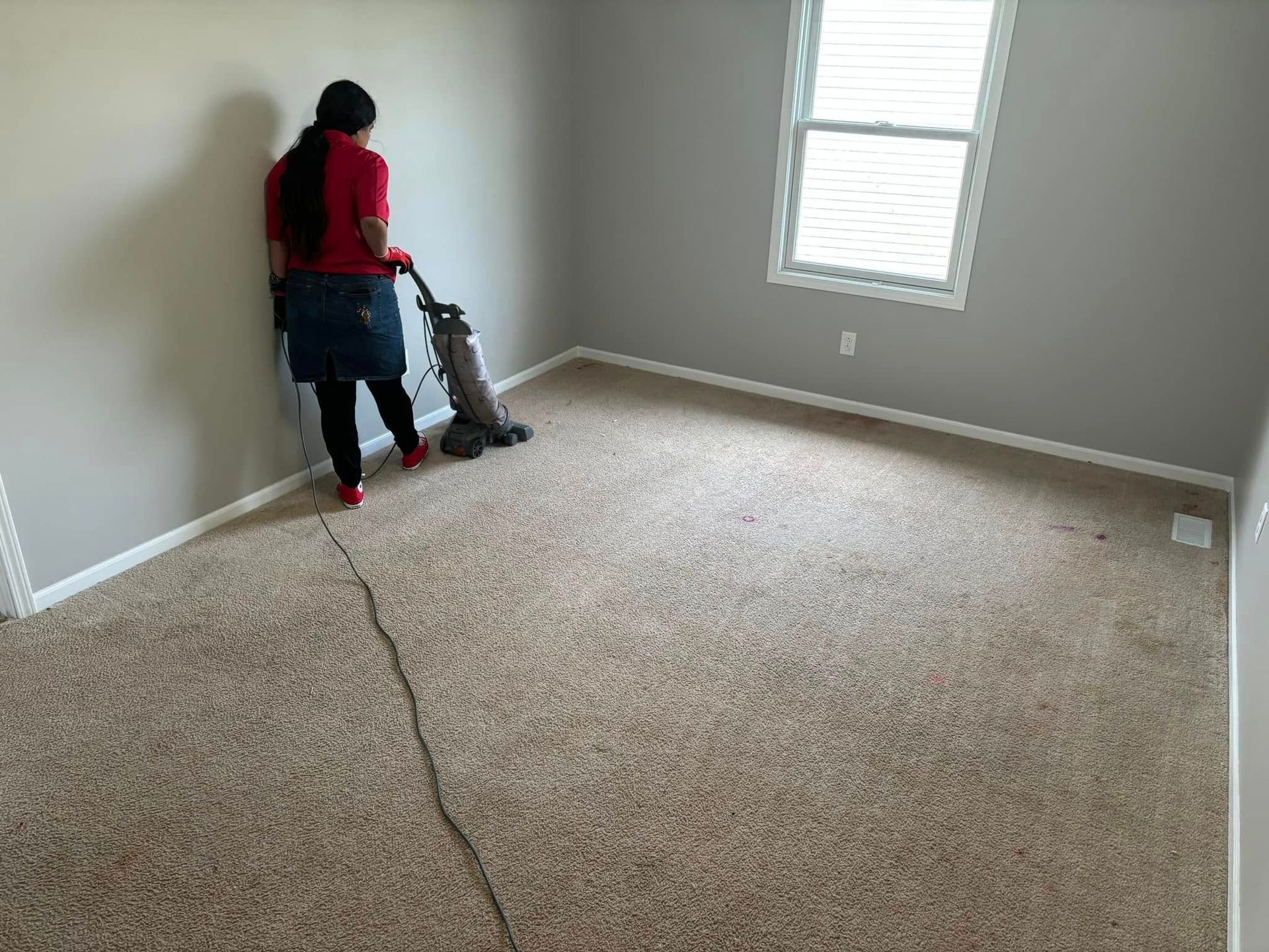 Person vacuuming beige carpet in a mostly empty room with a window and gray walls