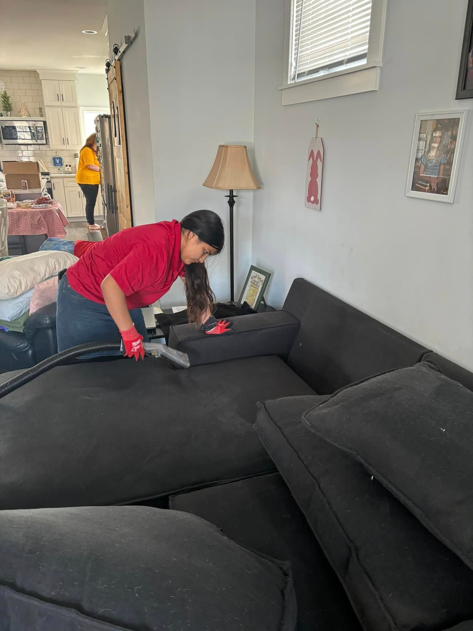 Person vacuuming a black sectional sofa in a living room with a lamp and wall art.