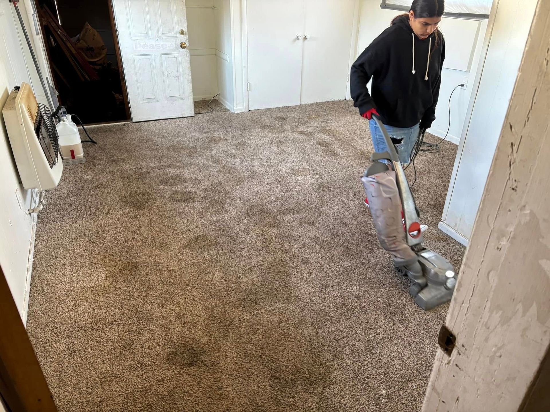Person vacuuming a beige carpet in an empty room with white walls and a closed door.