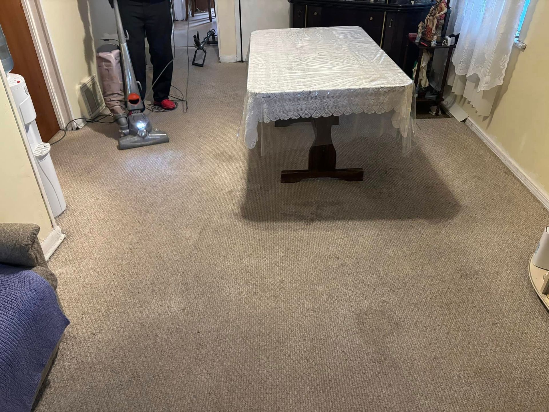 Carpeted living room with a table in the center and a person vacuuming near the doorway.