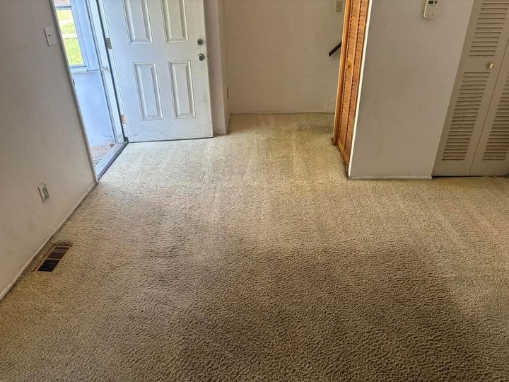 Empty carpeted hallway with white walls, open door, and vent near the floor.