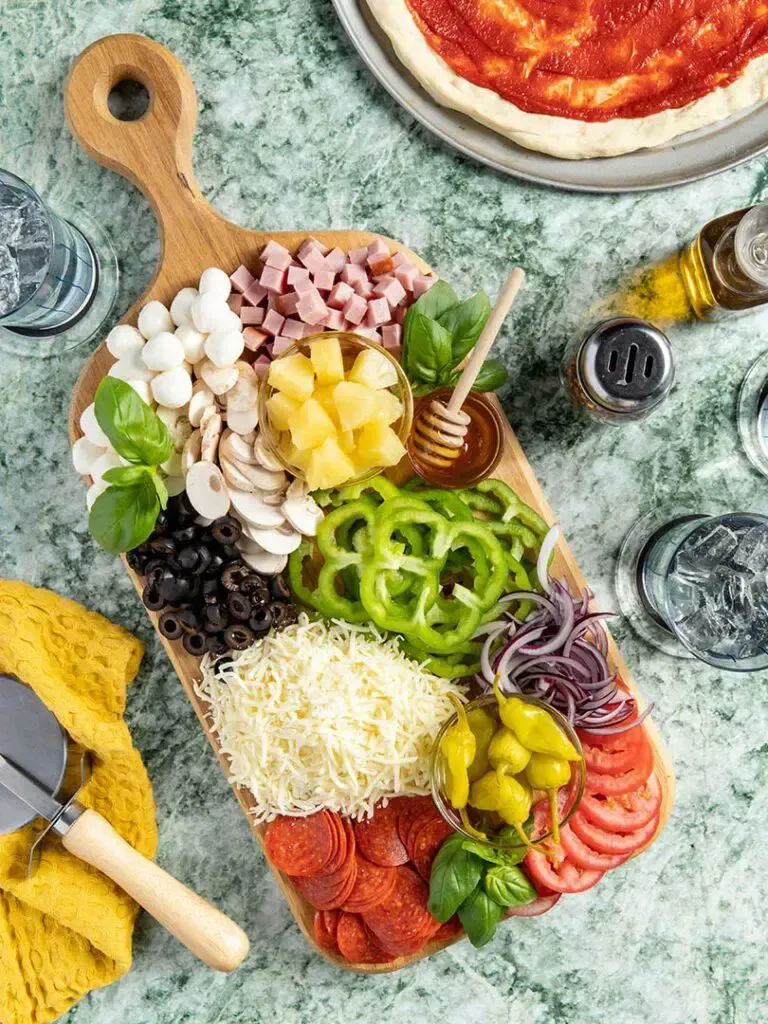 Pizza topping arrangement on a wooden board, with ingredients like olives, ham, peppers, and cheese.