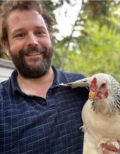 Man with a beard smiling, holding a white and gray chicken outdoors.