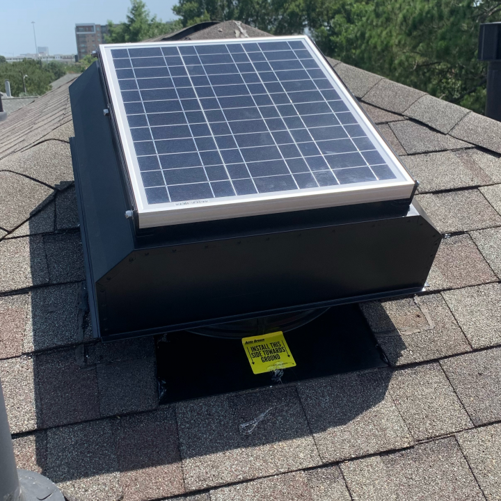 Solar-powered roof vent on a shingled roof. The vent is black with a rectangular solar panel on top.