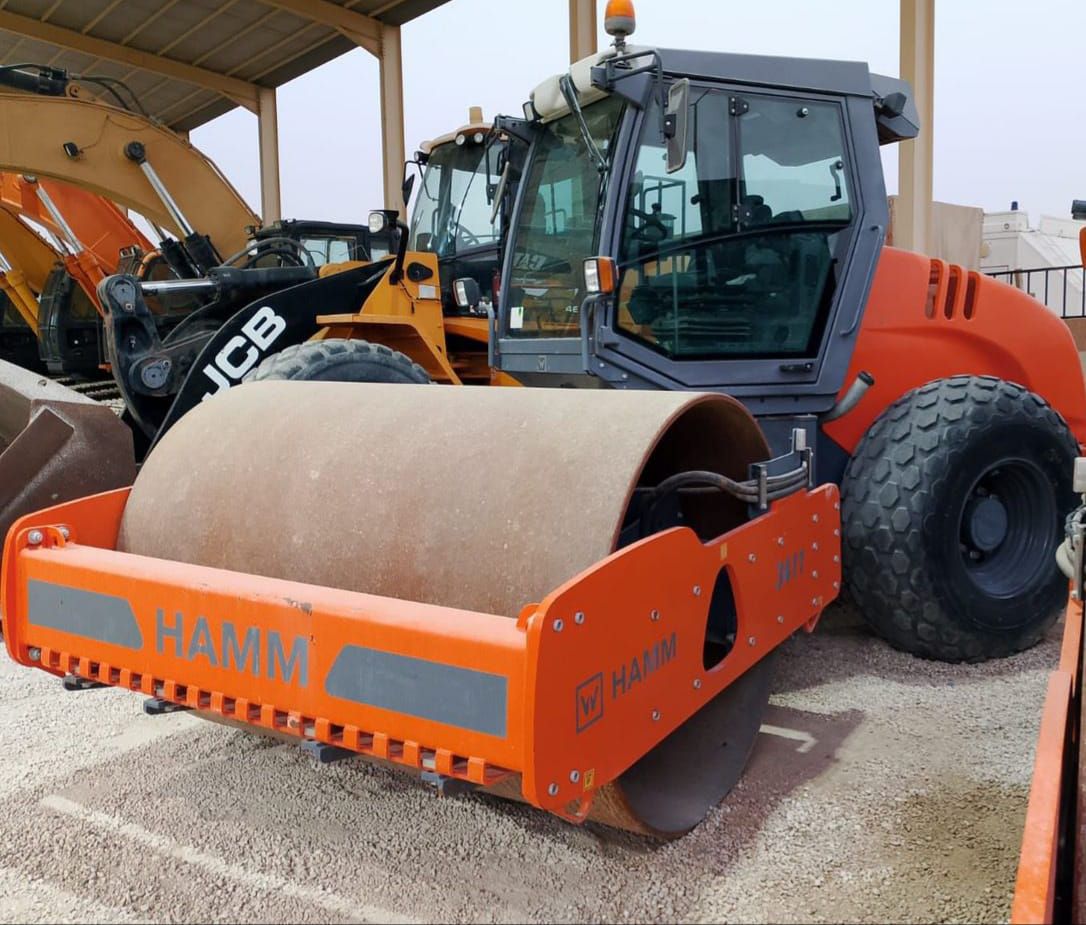 Orange and gray Hamm road roller on gravel, with JCB excavator in background.