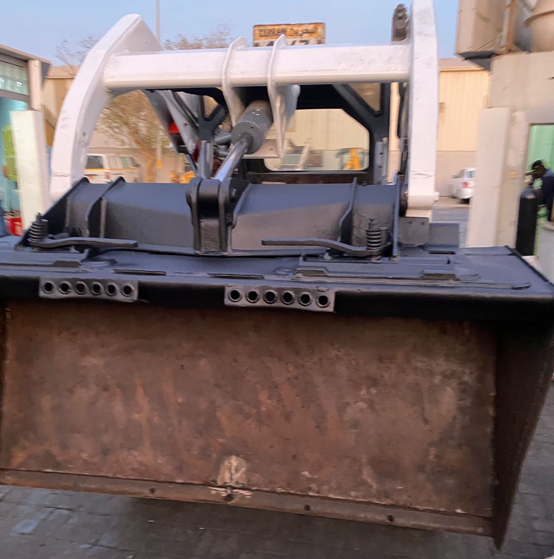 Front view of a black and white loader bucket and arm in a construction yard.