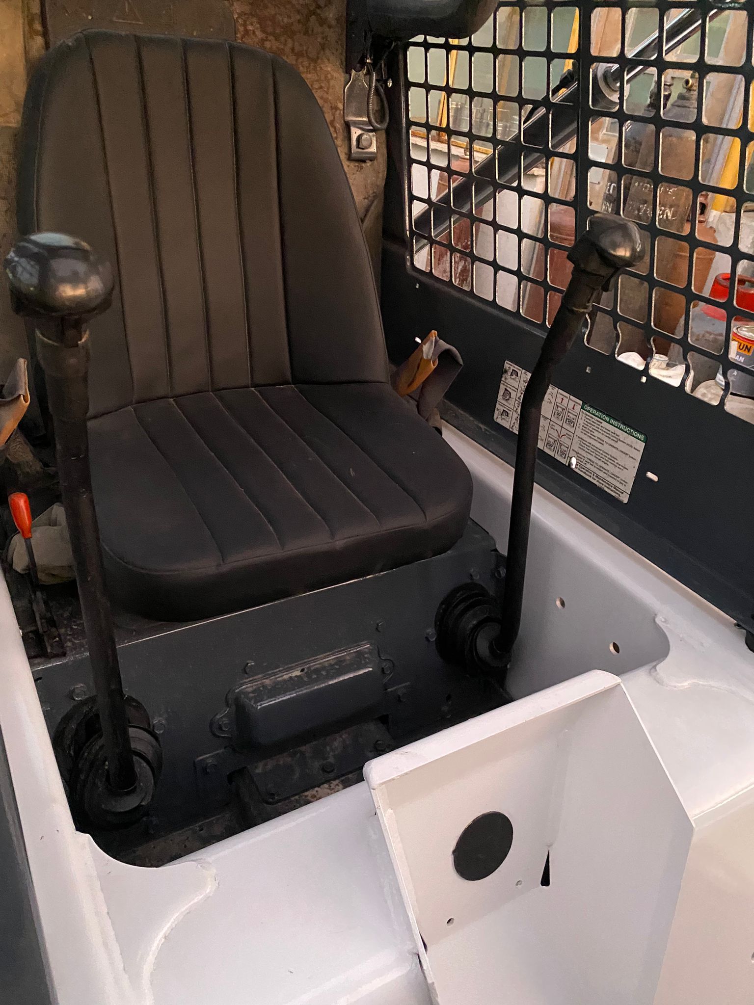 Interior view of a skid steer loader. Black seat and control levers against white and gray surfaces.