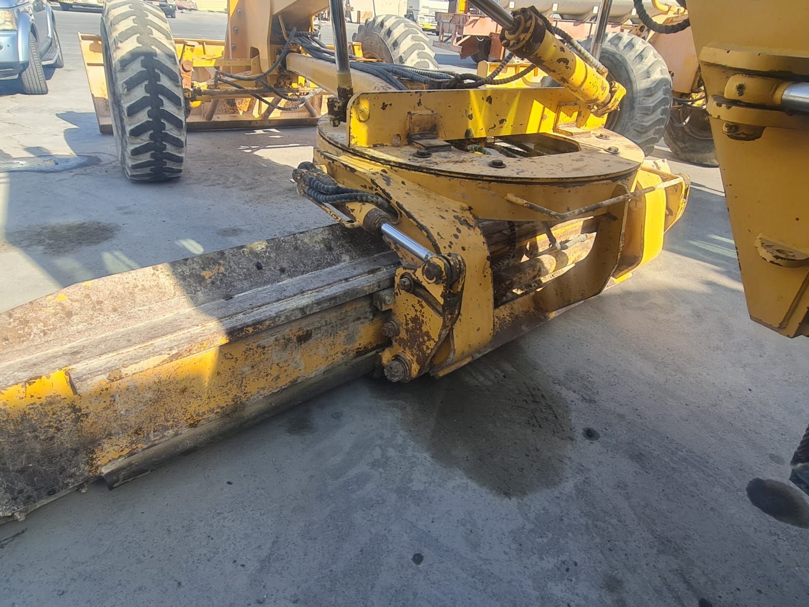 Yellow road grader blade angled down, outdoors on concrete.