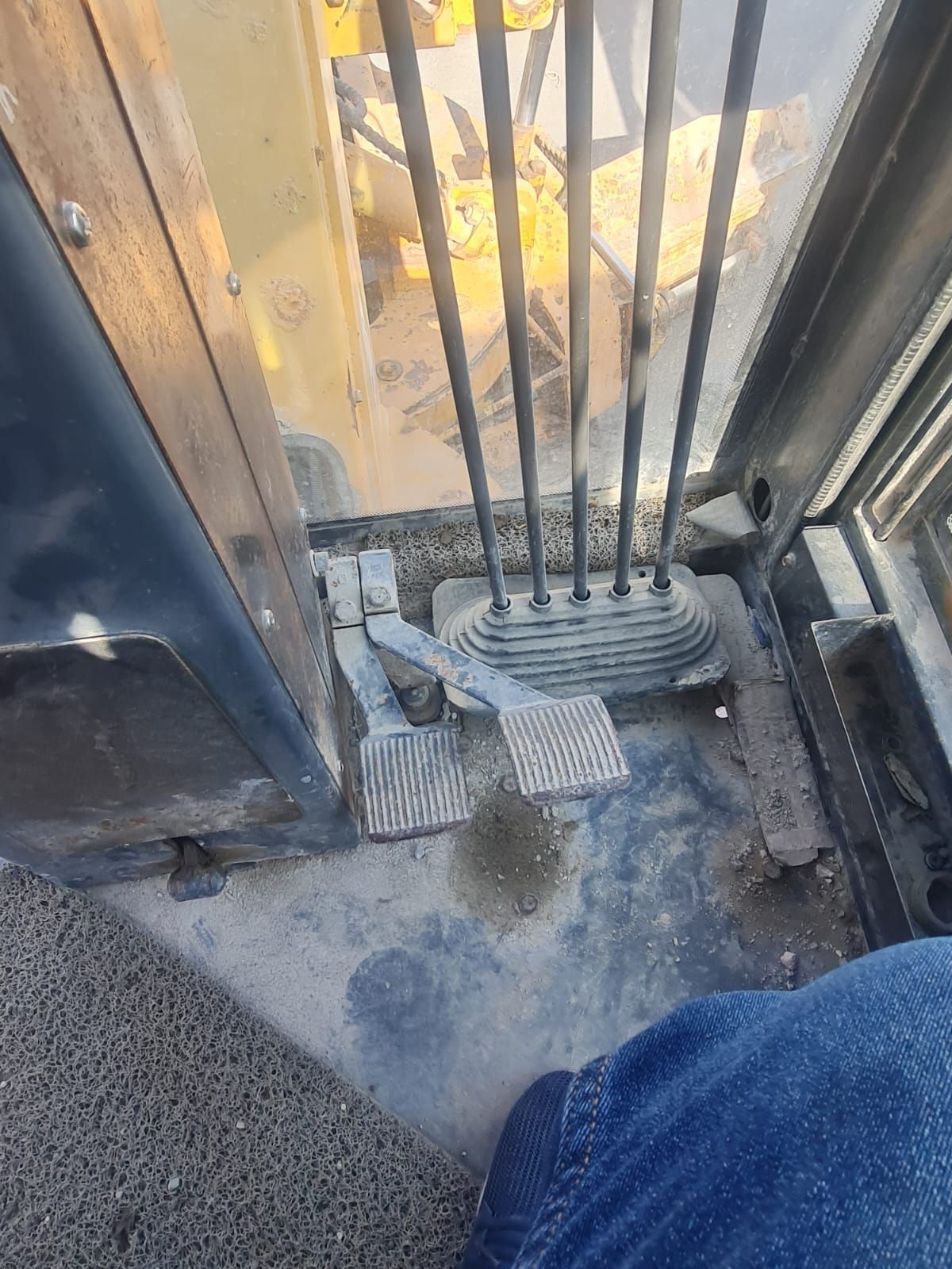 Inside a yellow excavator cab: levers, pedals, dirty floor. A person's leg in jeans is visible.