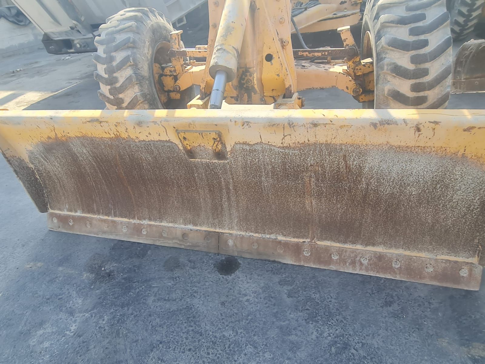 Yellow grader blade covered in dirt, with tires and machine visible.
