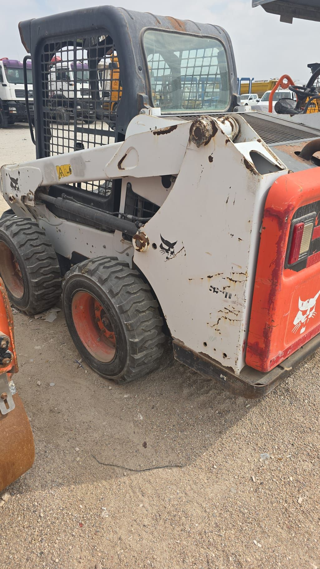 Bobcat skid-steer loader, weathered white and orange, on gravel. Black cab with cage.