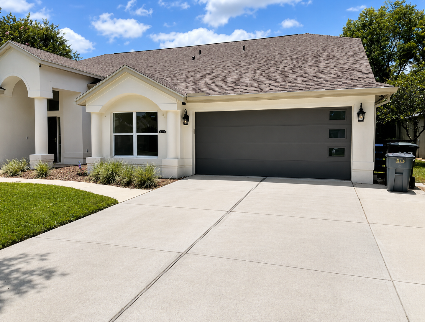 Suburban house with gray garage door, light stucco exterior, and a wide concrete driveway under a blue sky