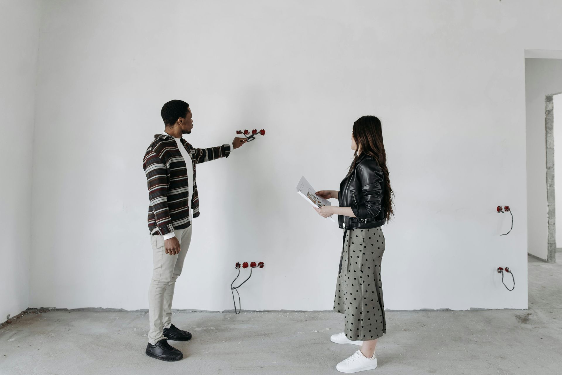 Two people in a bare room, one pointing at the wall while the other holds papers.