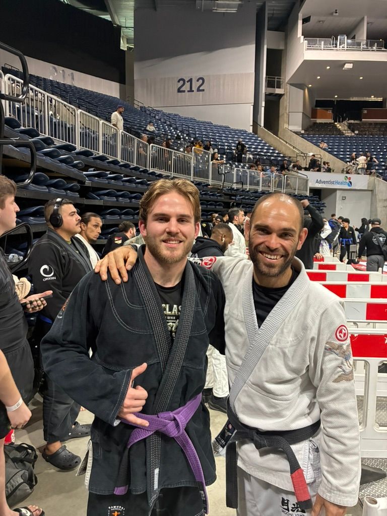 Two smiling people in martial arts gis posing in an indoor arena with bleachers behind them.