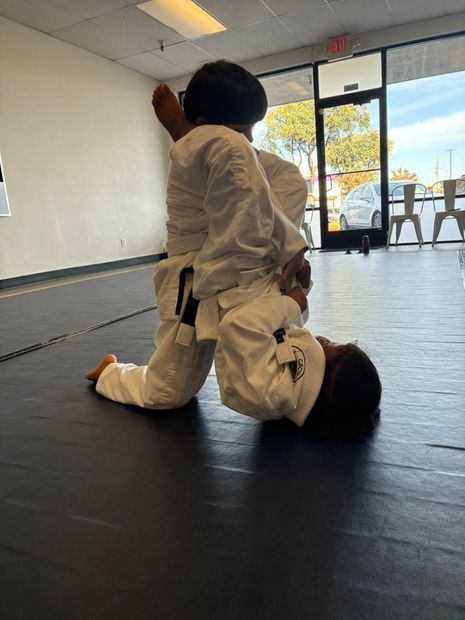 Two martial artists in white gi practice a grappling hold on black mats in a dojo