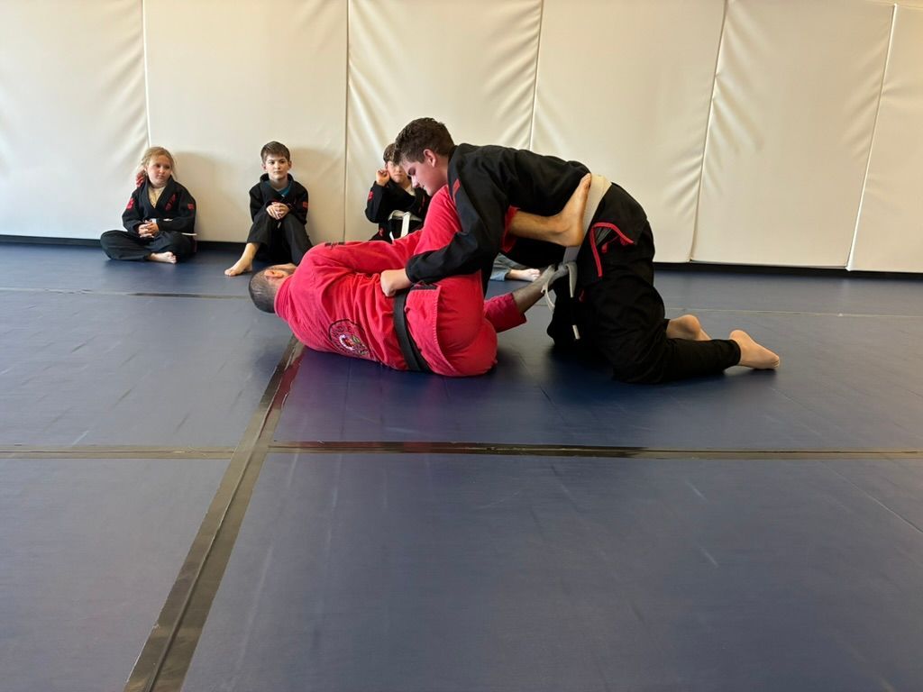 Two people grappling on blue mats in a dojo while two children watch from the side