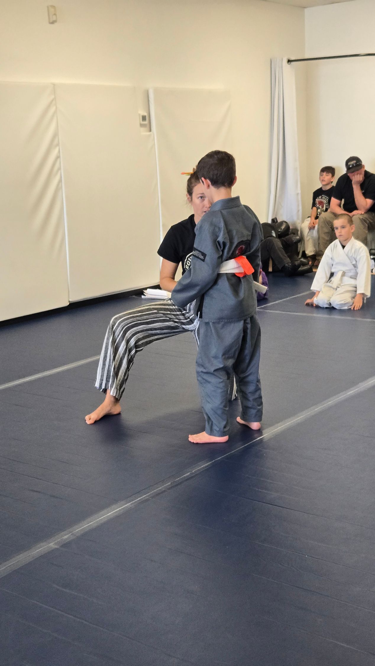 Two martial arts students practice a hold on a blue mat while others watch from the side.