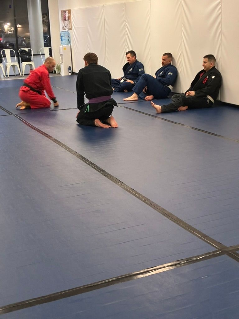 People seated on mats in a dojo, listening to an instructor in a red gi.