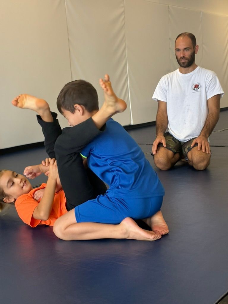Children practicing grappling on mats while an instructor watches in the background
