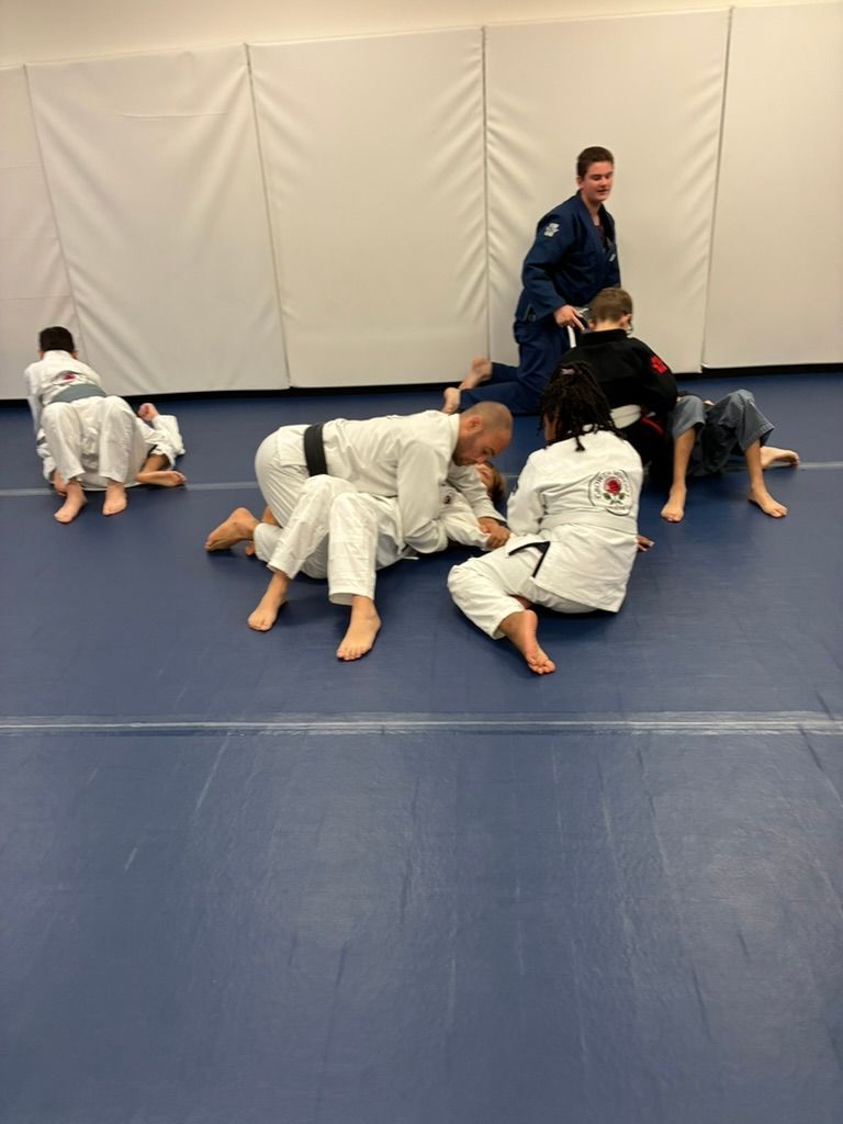 Children in white gis grapple on a blue mat while an instructor stands nearby in a dojo.