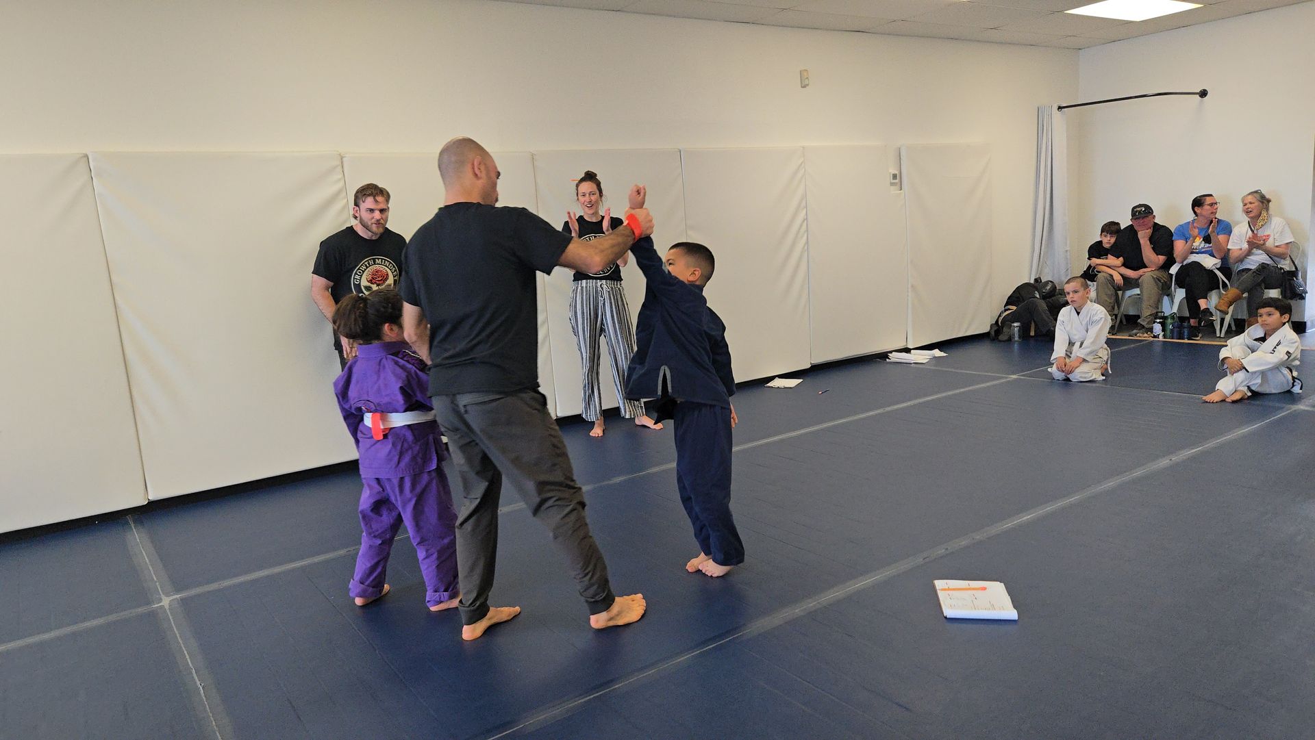 Children practicing martial arts with an instructor in a gym, spectators seated along the wall.