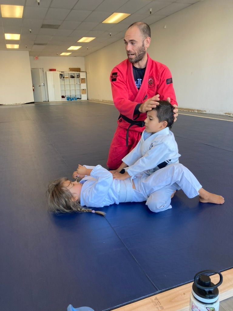 Instructor in red gi guiding a child in white gi during a grappling drill on blue mats.