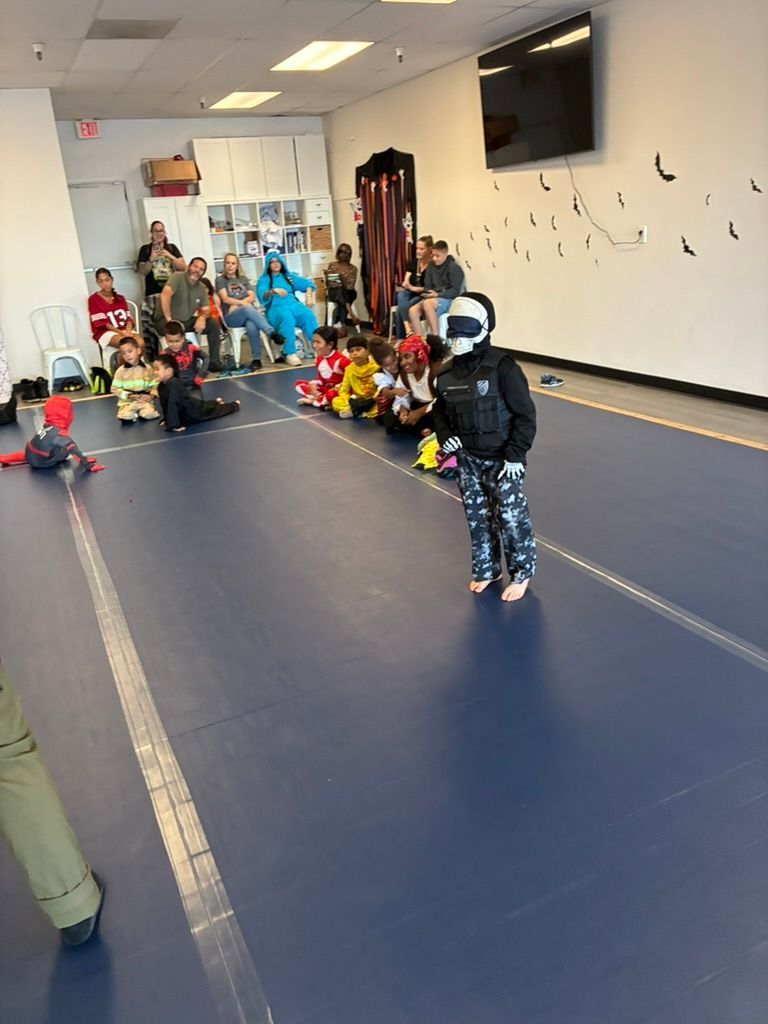 Children in a gym room with mats, toys, and adults seated along the back wall