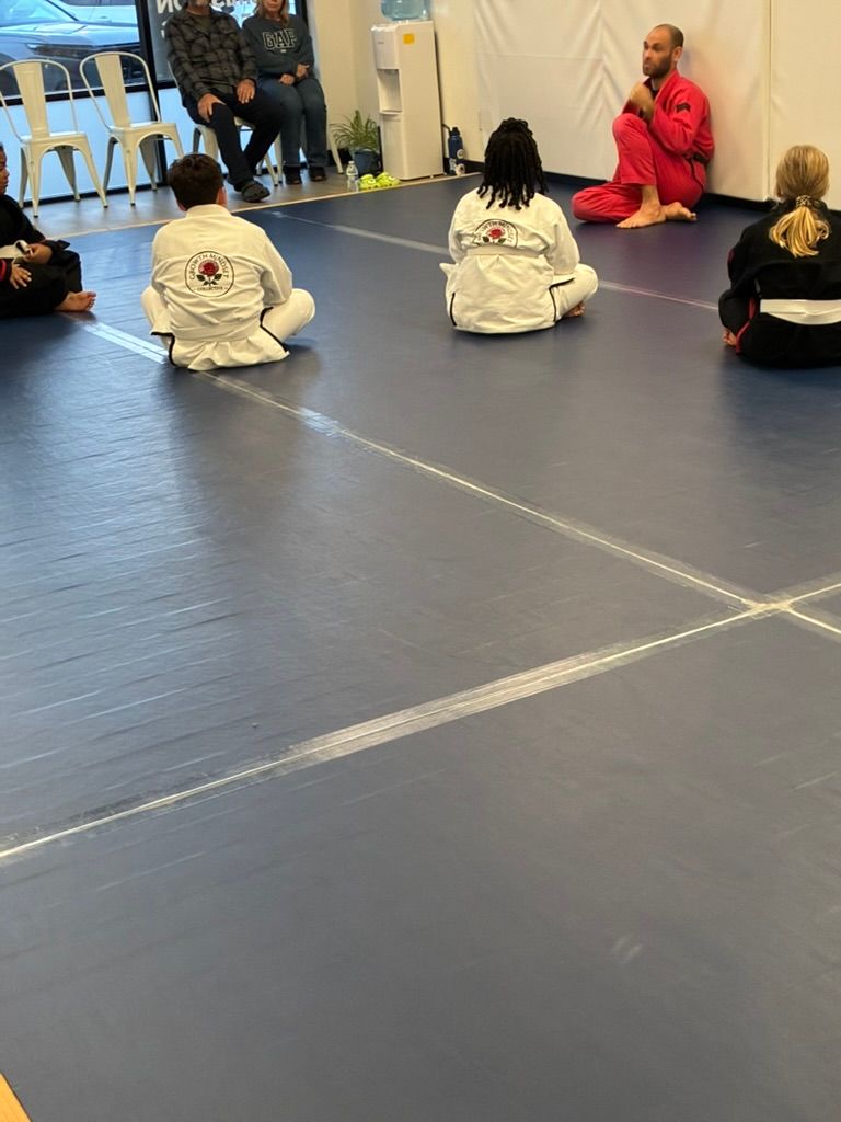 Children sitting on a blue mat in a dojo, facing an instructor in a red shirt.