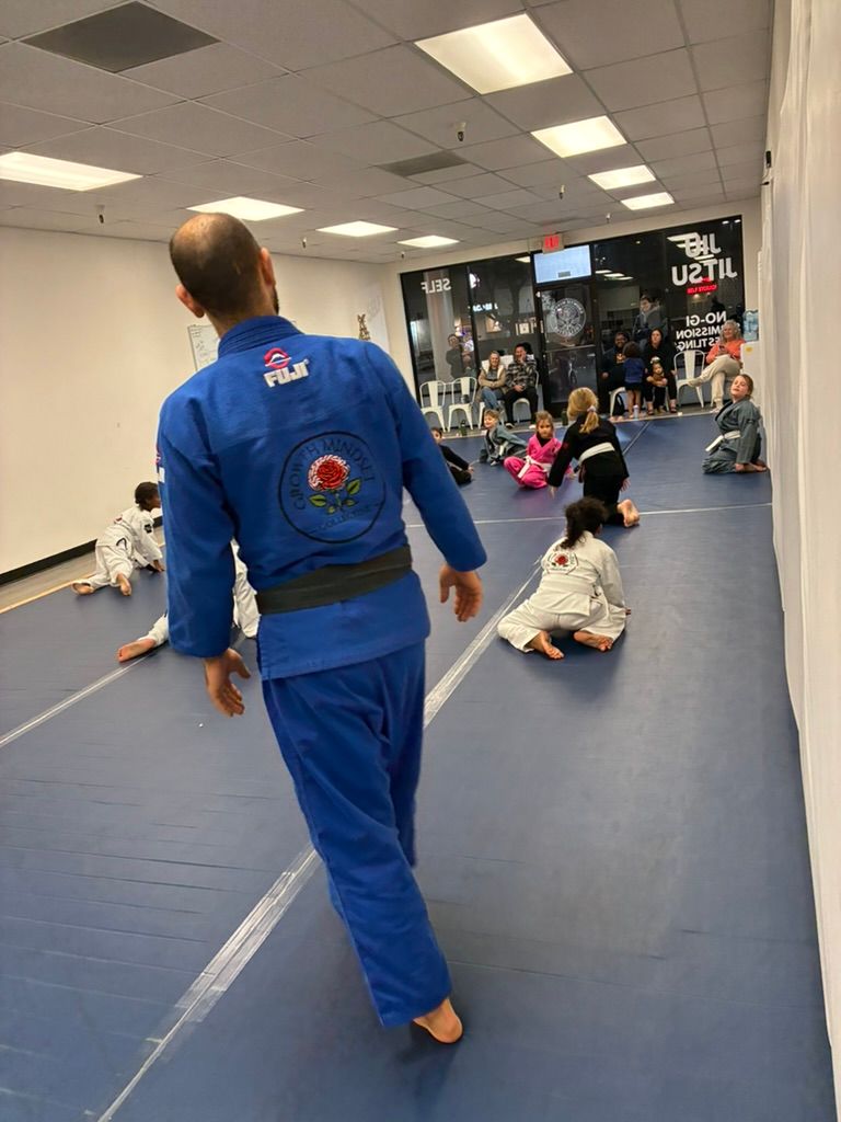 Martial arts class on blue mats, instructor in blue gi leading seated students in a studio
