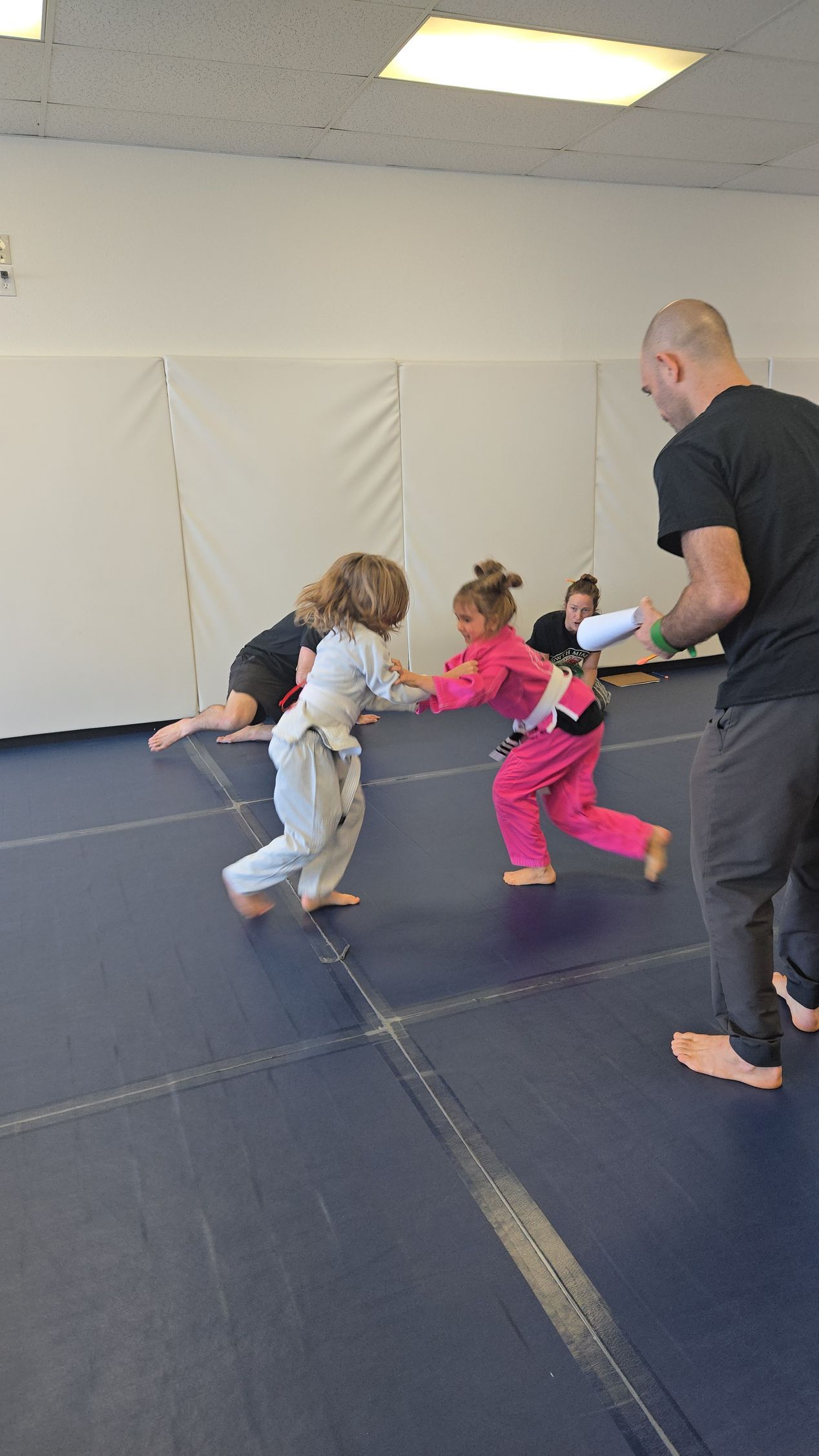 Children in colorful outfits practice martial arts on mats while an instructor watches nearby.