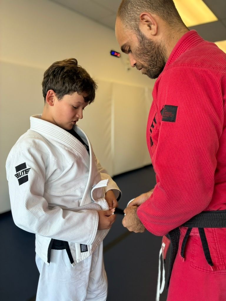 Two people in white and red martial arts gis adjusting a belt in a training room