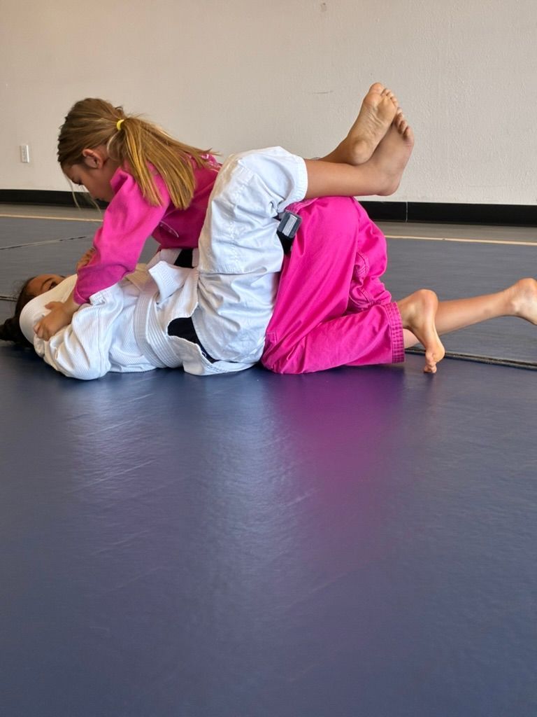 Two children practicing grappling on a mat, one in a pink gi pinning the other in white and pink.