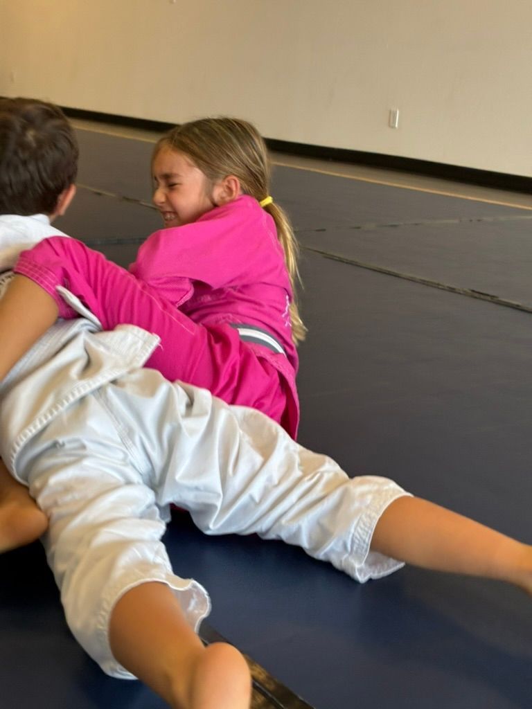 Two children grappling on a mat, one in pink and one in white, in a gym setting