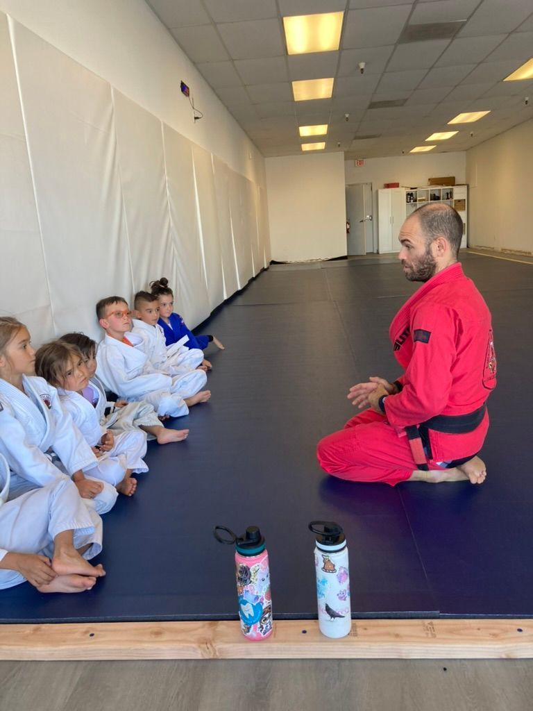 Martial arts instructor in red kneels before a line of children in white gis on a mat in a bright studio