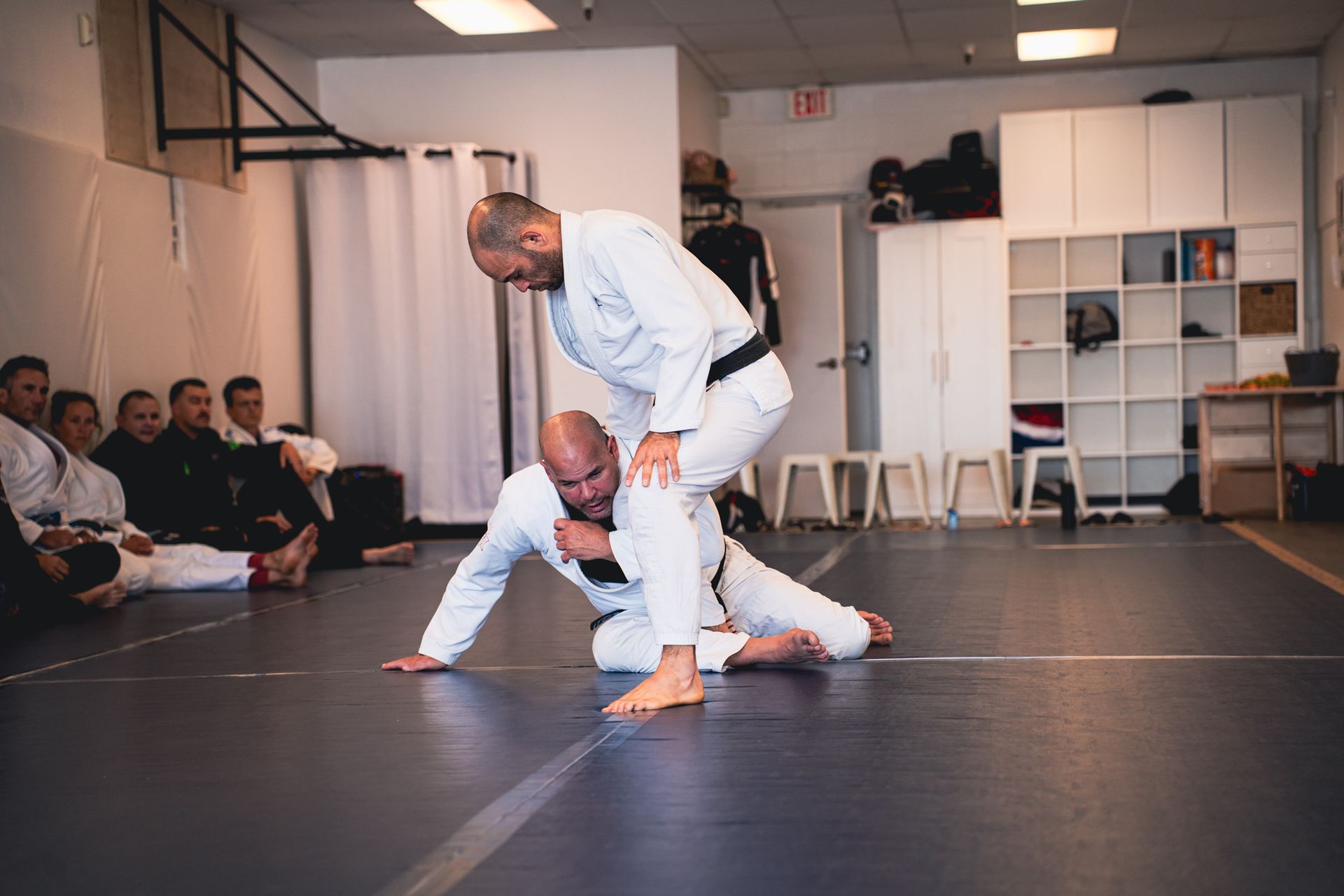 Two people practicing martial arts on black mats in a training studio, with spectators seated along the wall.